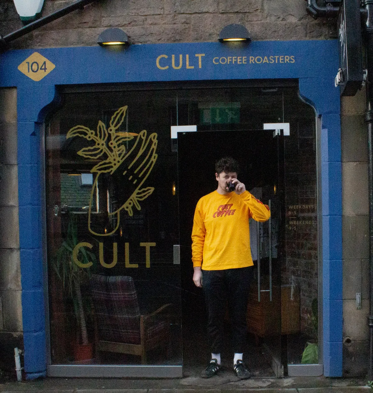 The blue shopfront of Cult Coffee Roasters in Newington, with a barista in a yellow Cult Coffee shirt in the doorway