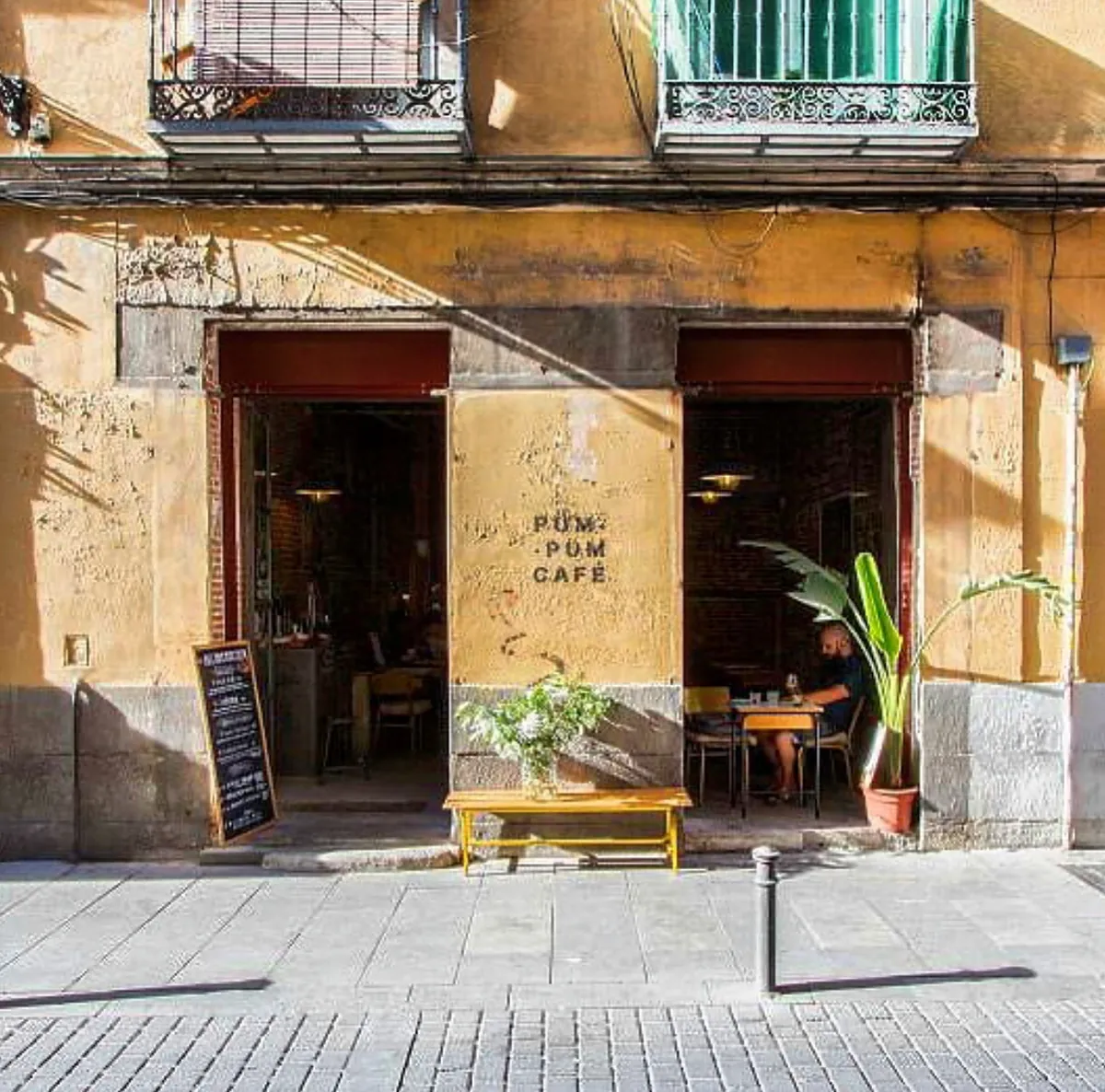 The yellow façade of Pum Pum Café in Lavapiés, with a bench and plant outside and a customer at a window table