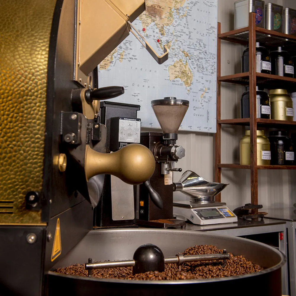 The brass and black drum roaster at ManCoCo with roasted beans cooling in the tray and a world map on the wall