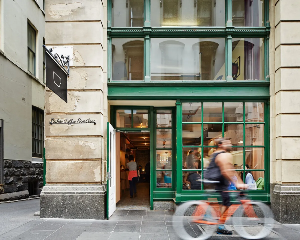 The green-framed shopfront of Dukes Coffee at Ross House, with a cyclist passing in front