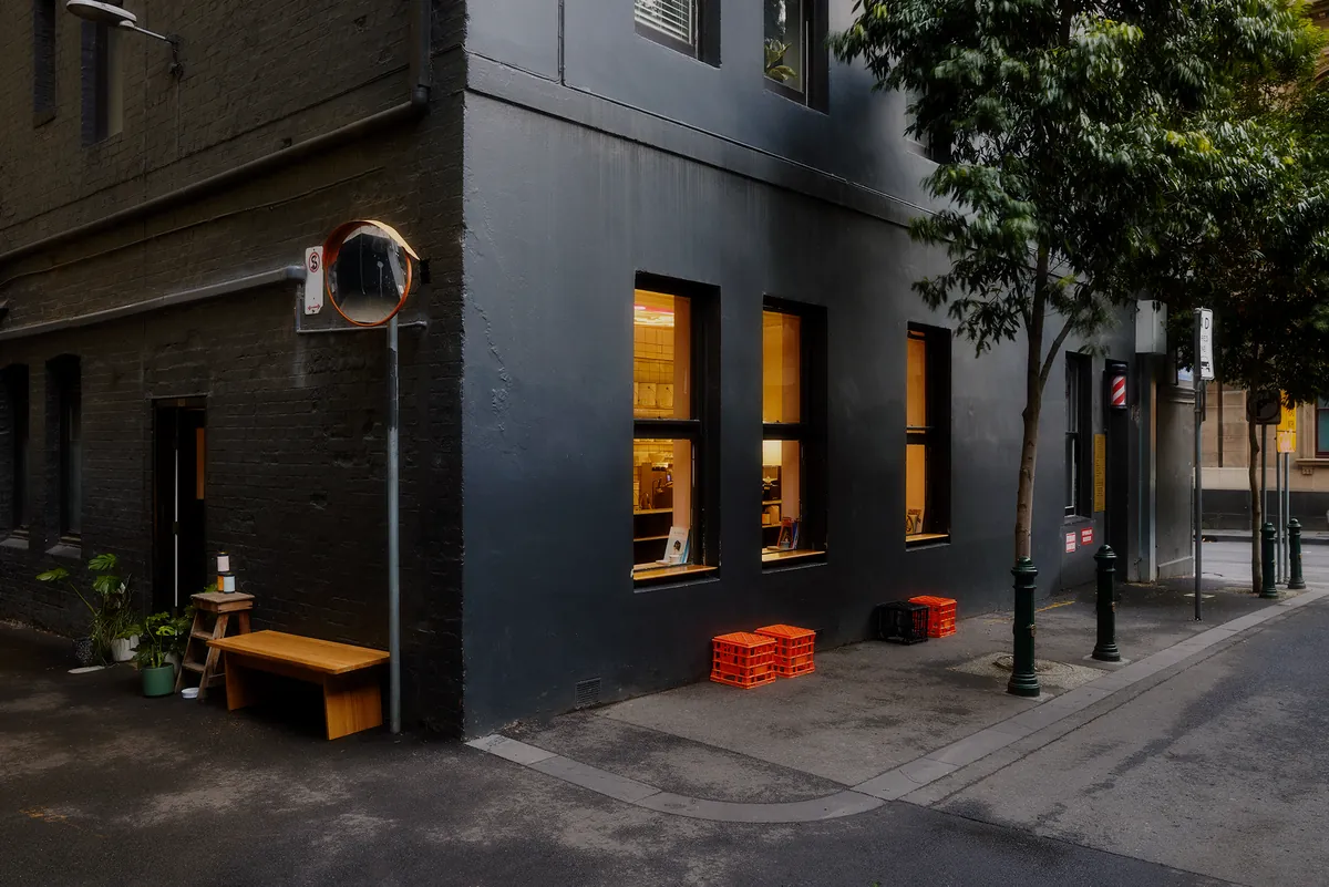 The black-painted corner of Patricia Coffee Brewers at dusk, with lit windows, milk crates on the pavement and a bench by the door