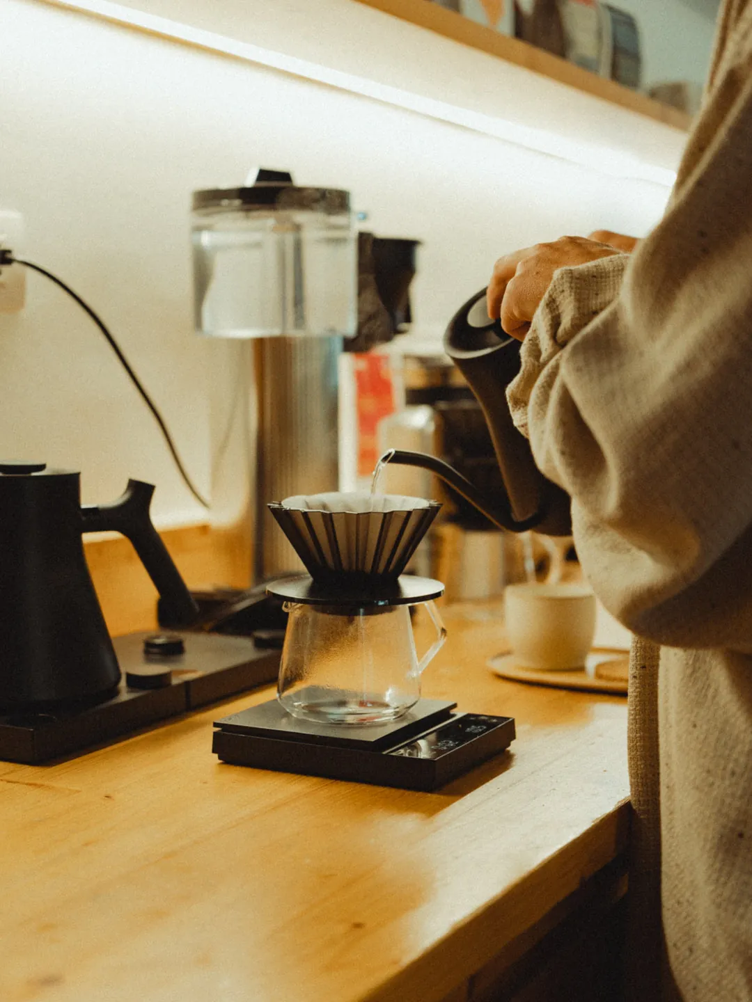 A barista pouring a V60 at Orsonero, with a kettle, scale and glass server on the wooden counter