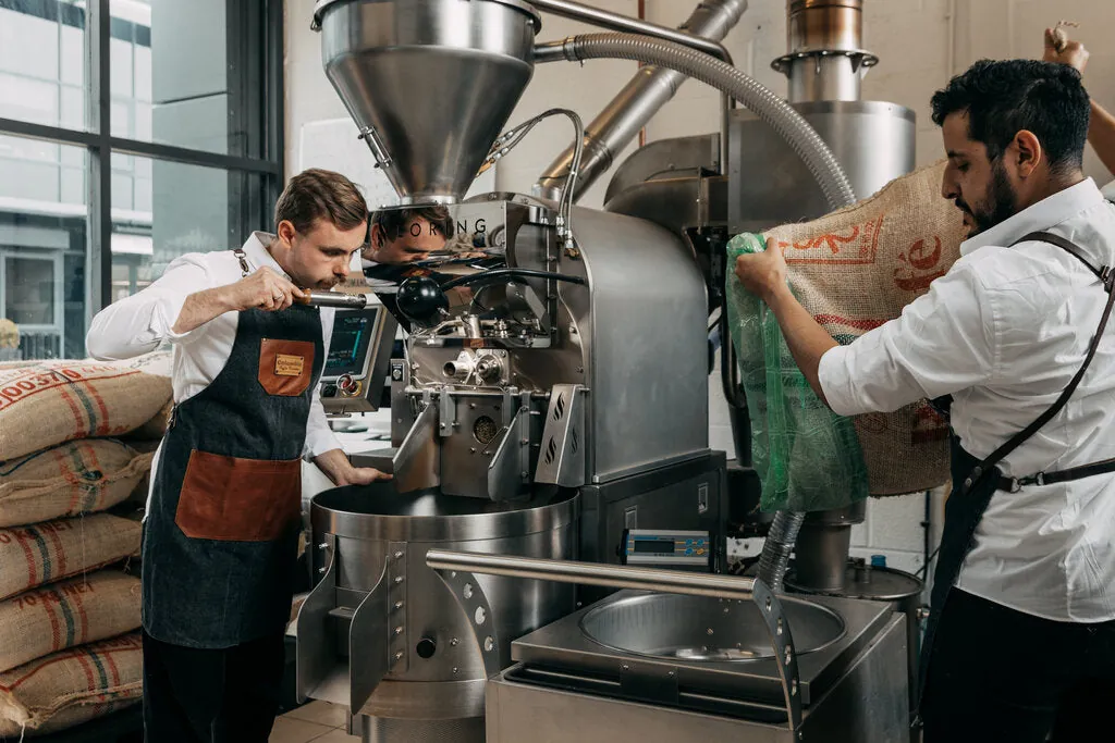 Two staff at work beside the coffee roaster at the Colombia Coffee Roasters roastery, with sacks of green coffee stacked by the window