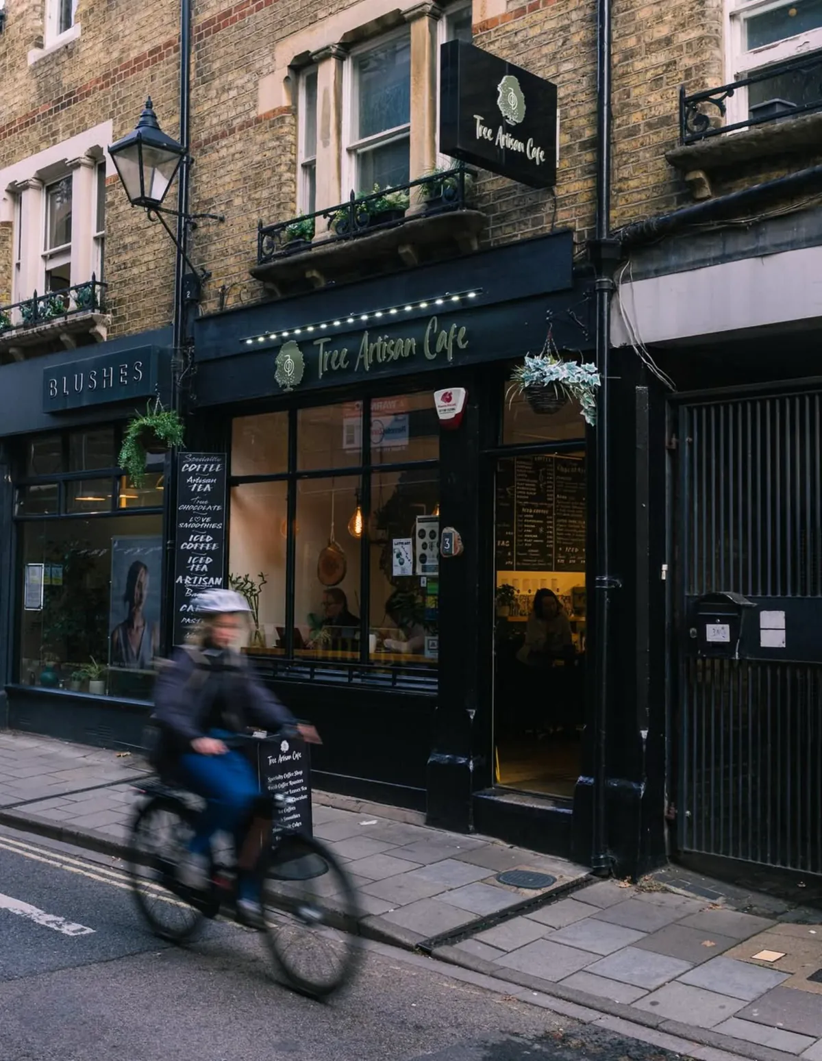 The dark green shopfront of Tree Artisan Cafe in Oxford, with its hanging sign and a cyclist passing