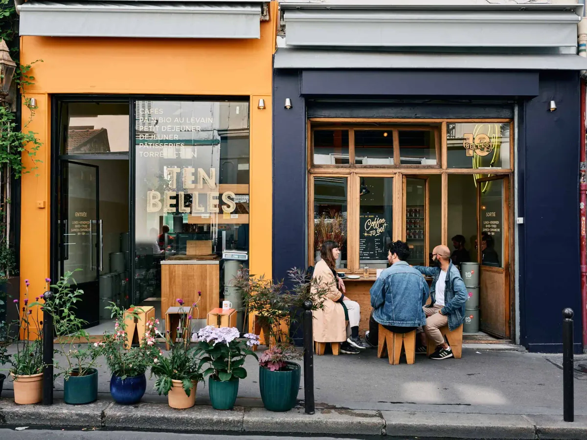 The yellow and navy frontage of Ten Belles near the Canal Saint-Martin, with three customers at the window counter and potted plants on the pavement