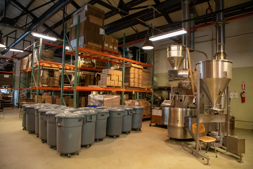 Inside the Brooklyn Roasting Company roastery, with grey bins, a roaster and racks of green coffee under a steel roof