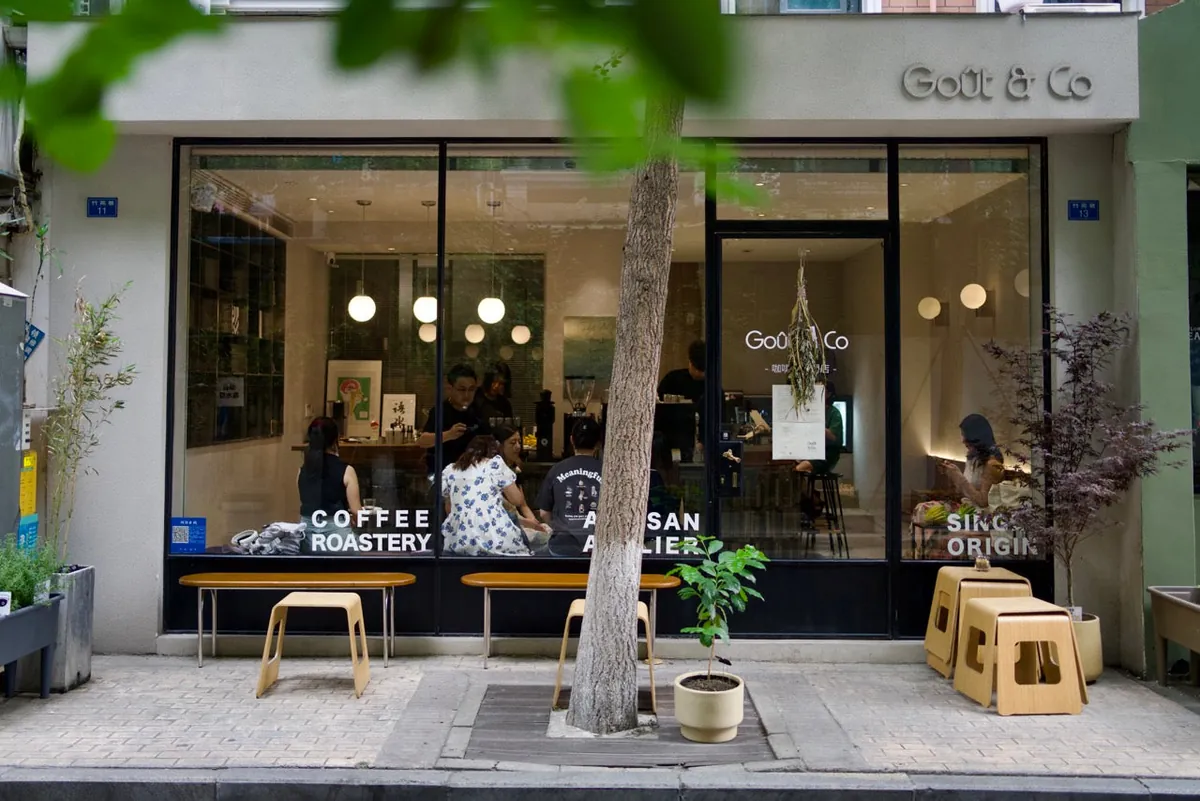 The glass shopfront of a Goût & Co café in Chengdu, with wooden benches and stools on the pavement under a tree