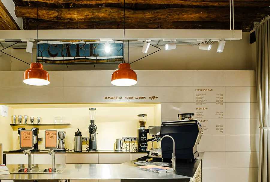 The espresso bar of the renovated Cafés El Magnífico shop in El Born, with orange pendant lamps under a timber-beamed ceiling