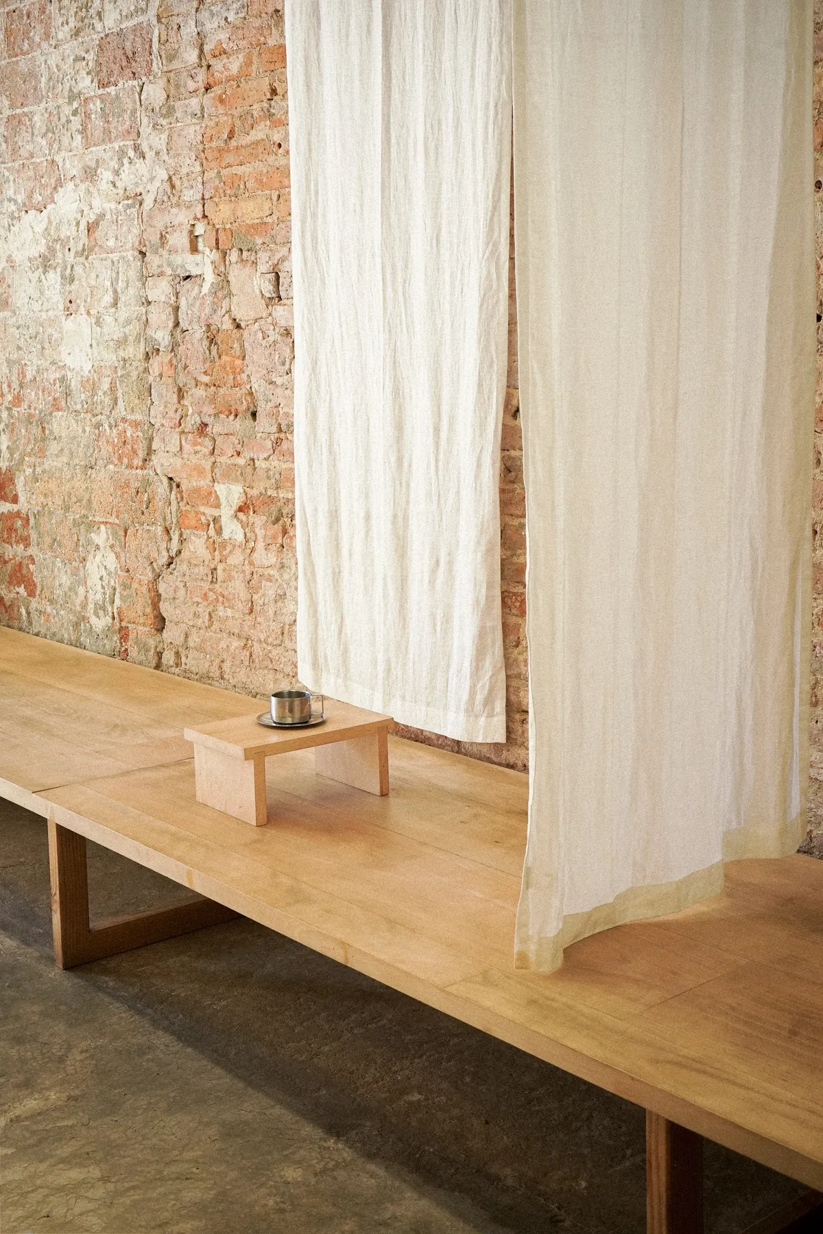 A long wooden bench against a bare brick wall inside Otsu, with white fabric curtains hanging over it