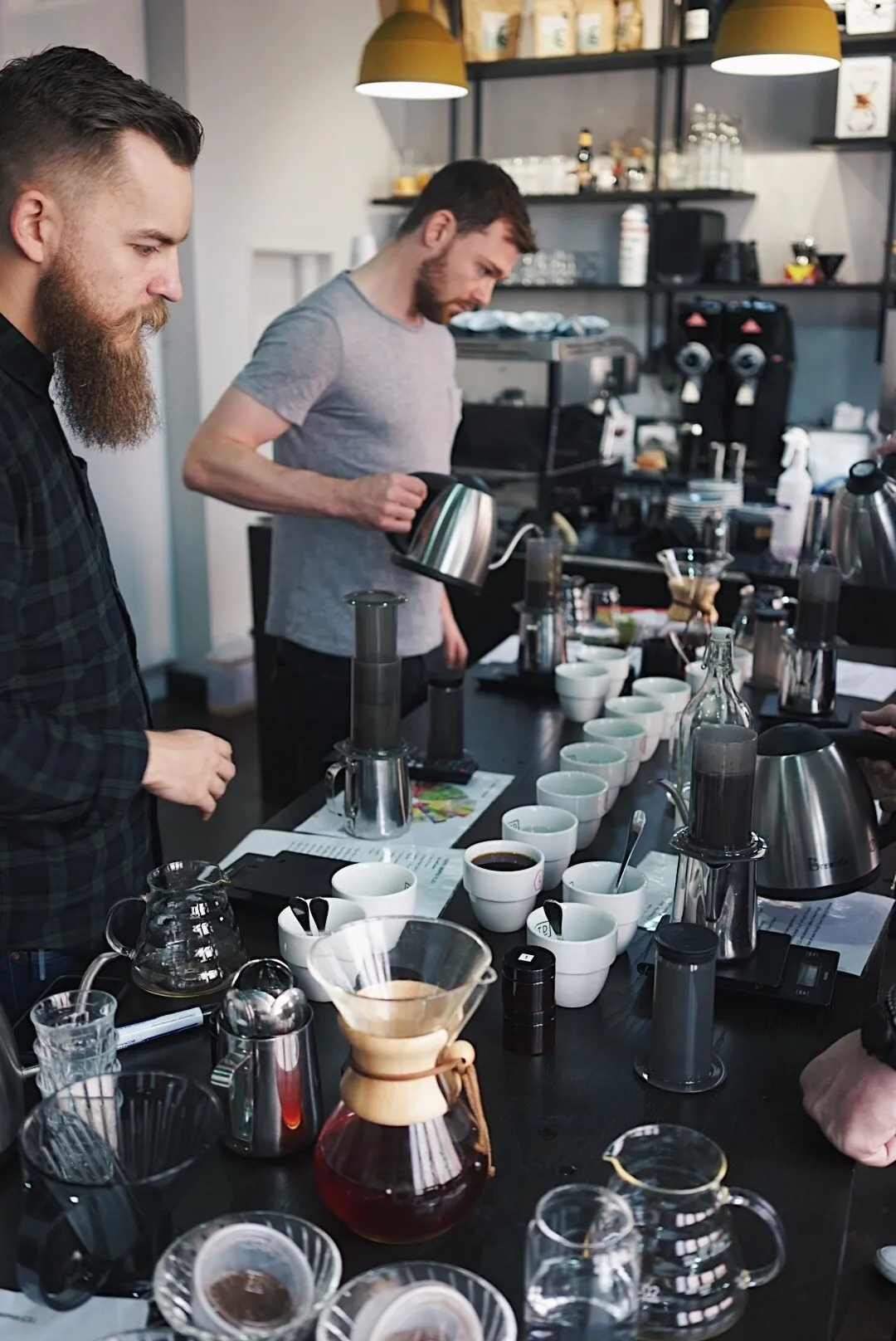 Two baristas pour filter coffee at a long brew bar laid with cupping bowls, AeroPresses and a Chemex during a brewing class at Established Coffee in Belfast