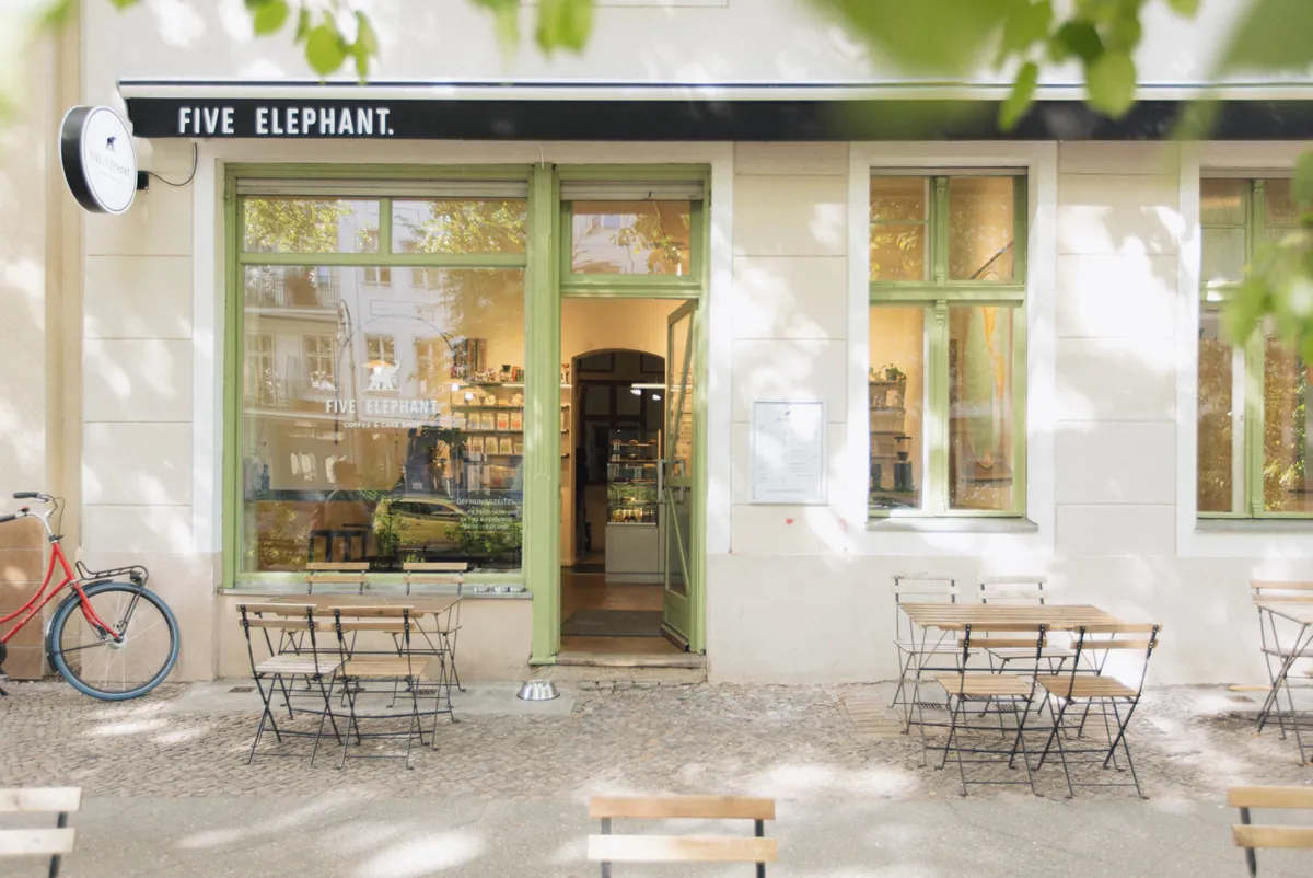 The Five Elephant café in Kreuzberg, with green-framed windows, an open door and folding tables on the pavement