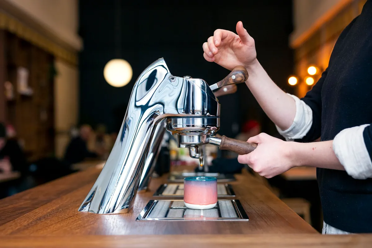 A barista working the chrome lever espresso machine on the wooden bar at Fortitude Coffee, with the café room softly lit behind
