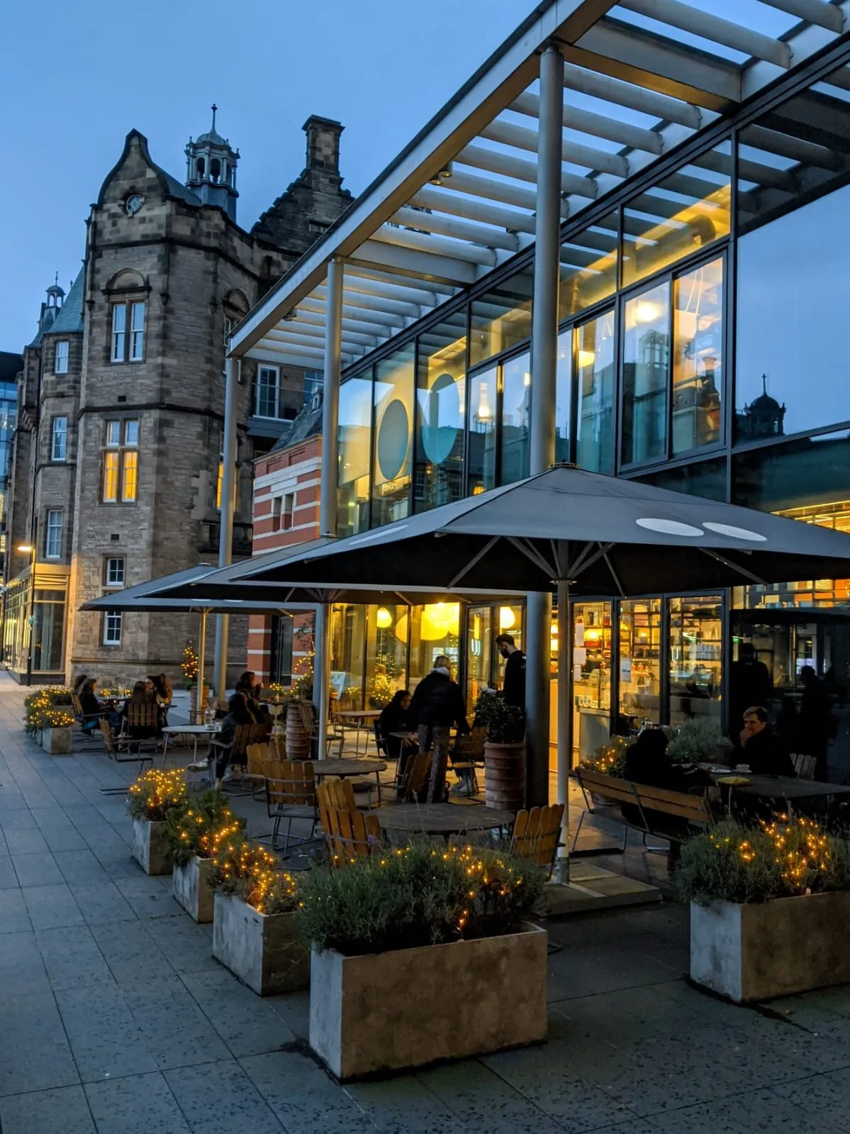 The glass-fronted Söderberg Pavilion Café at Quartermile at dusk, with planters strung with lights and people at tables under parasols