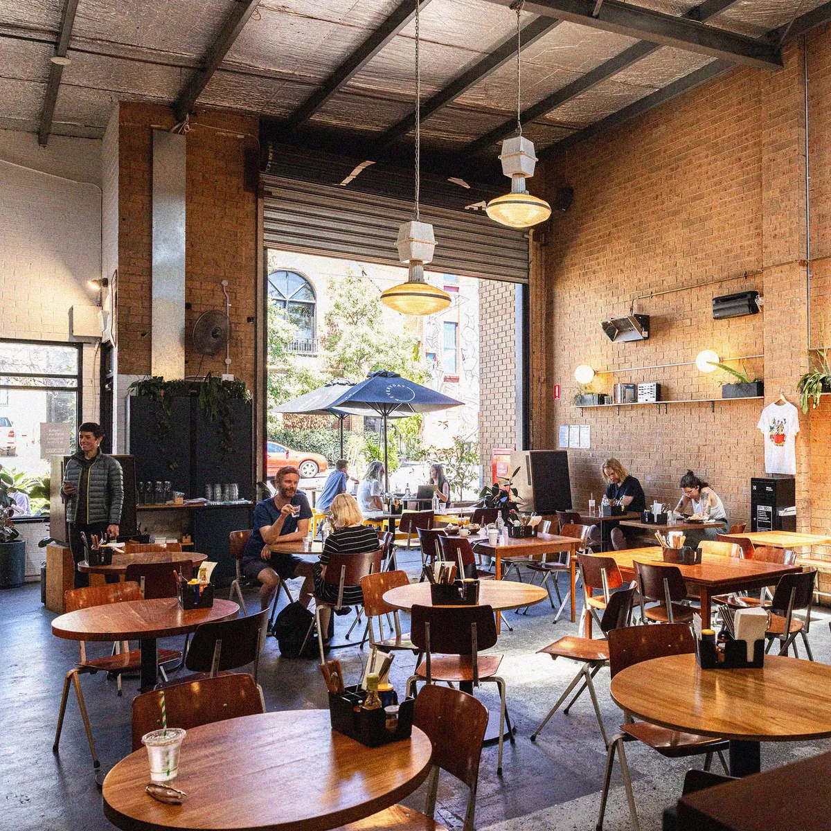 The high-ceilinged warehouse room at Everyday Coffee in Collingwood, with round timber tables, brick walls and an open roller door onto the street