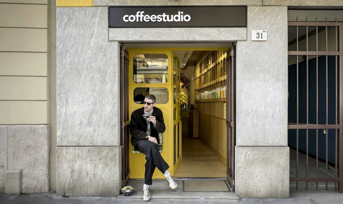 The narrow yellow doorway of the Sevengrams coffee studio in Porta Venezia, under a black coffeestudio sign, with a man seated on the step