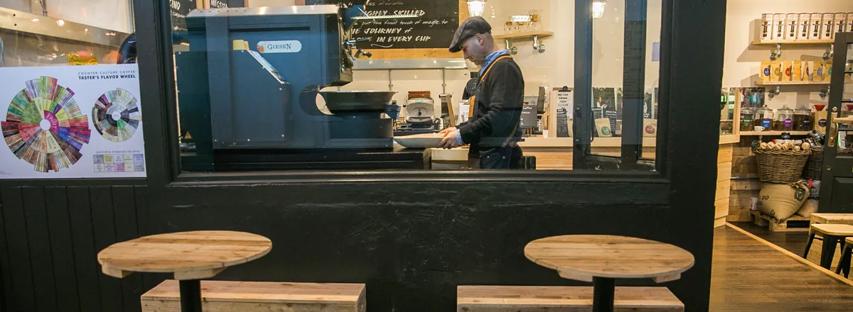 The glass front of a Colombia Coffee Roasters café, with a barista at the espresso machine behind the window and wooden stools outside