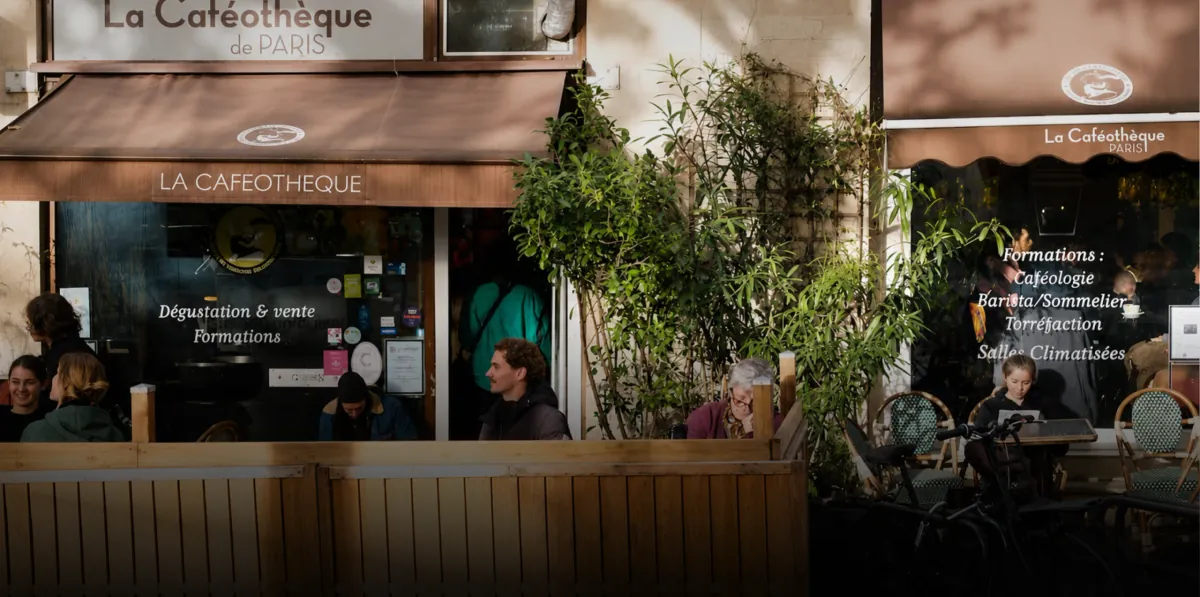 The awning-covered frontage of La Caféothèque, with people seated on its fenced terrace among potted plants