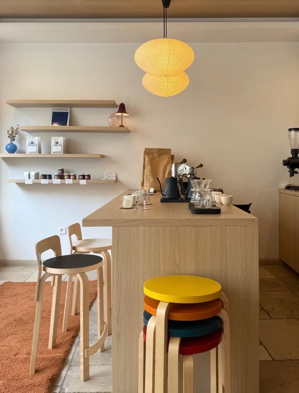 The blond-wood bar at Klover Coffee Paris, with stacked coloured stools, shelves of coffee bags and a paper pendant lamp