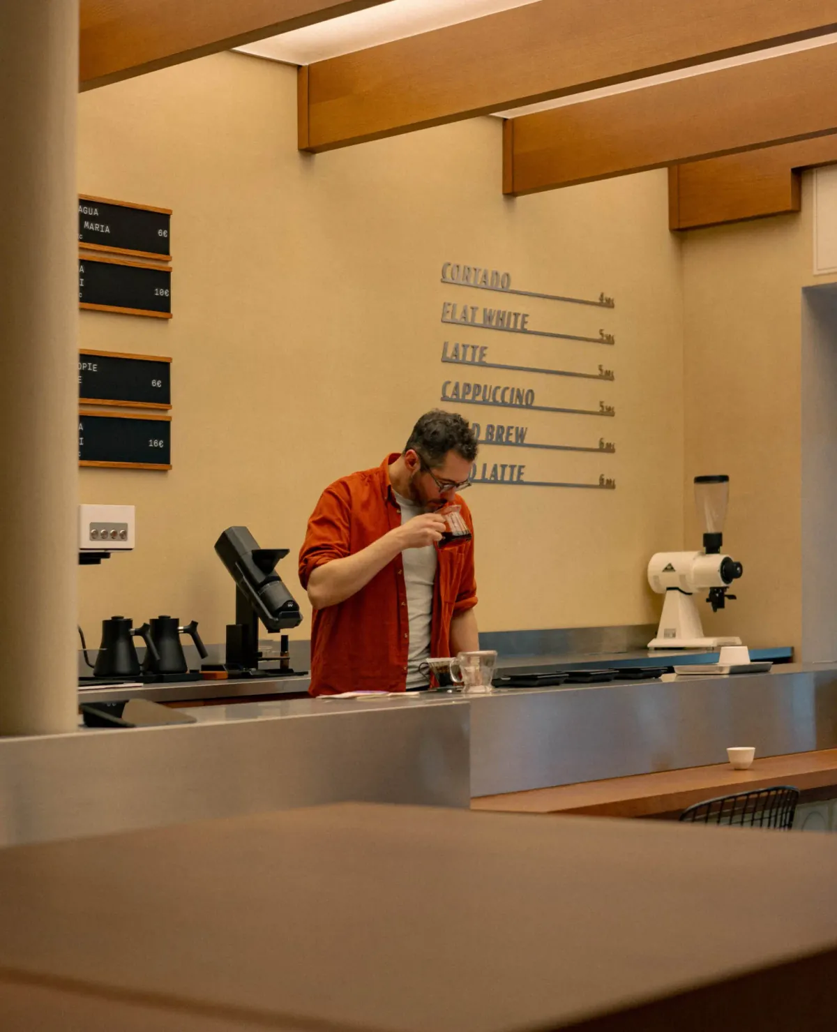 A barista at work behind the steel counter of a Tanat coffee shop, with the drinks menu lettered on the wall