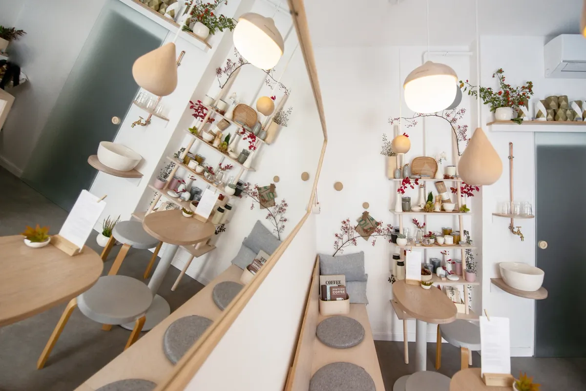 The small white room at 4minutes cafe, with a long wooden bench, round wooden tables, pale wooden pendant lamps and shelves of ceramics and berry branches reflected in a large mirror