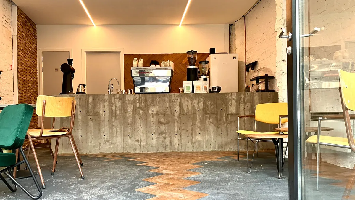 The Astrid bar of Full Circle Coffee, a board-marked concrete counter with an espresso machine, grinders and retail coffee bags, a herringbone wood strip across a grey floor and yellow and green chairs