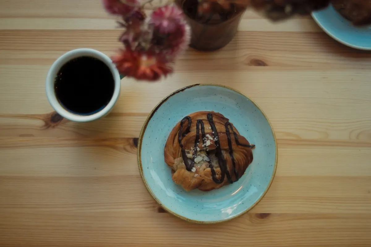 A cup of black filter coffee and a chocolate-drizzled sourdough pastry on a blue plate on a wooden table at Kaf