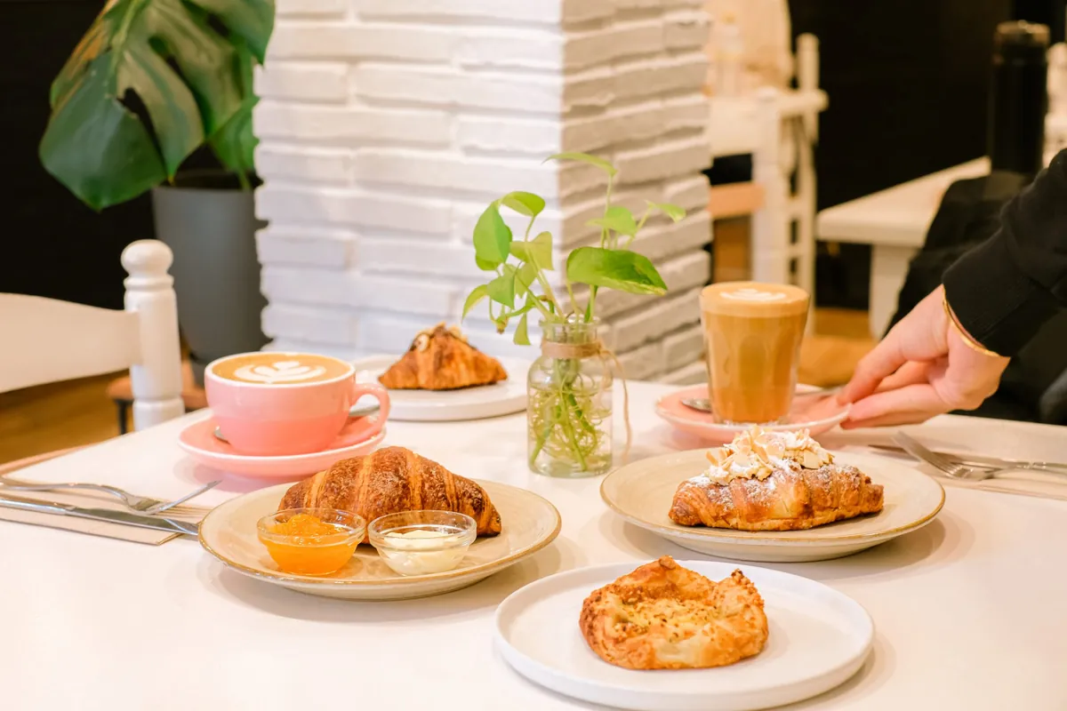 A table at Let It Bloom with croissants, a pastry, a latte in a pink cup and a glass of coffee in front of a white brick pillar and plants