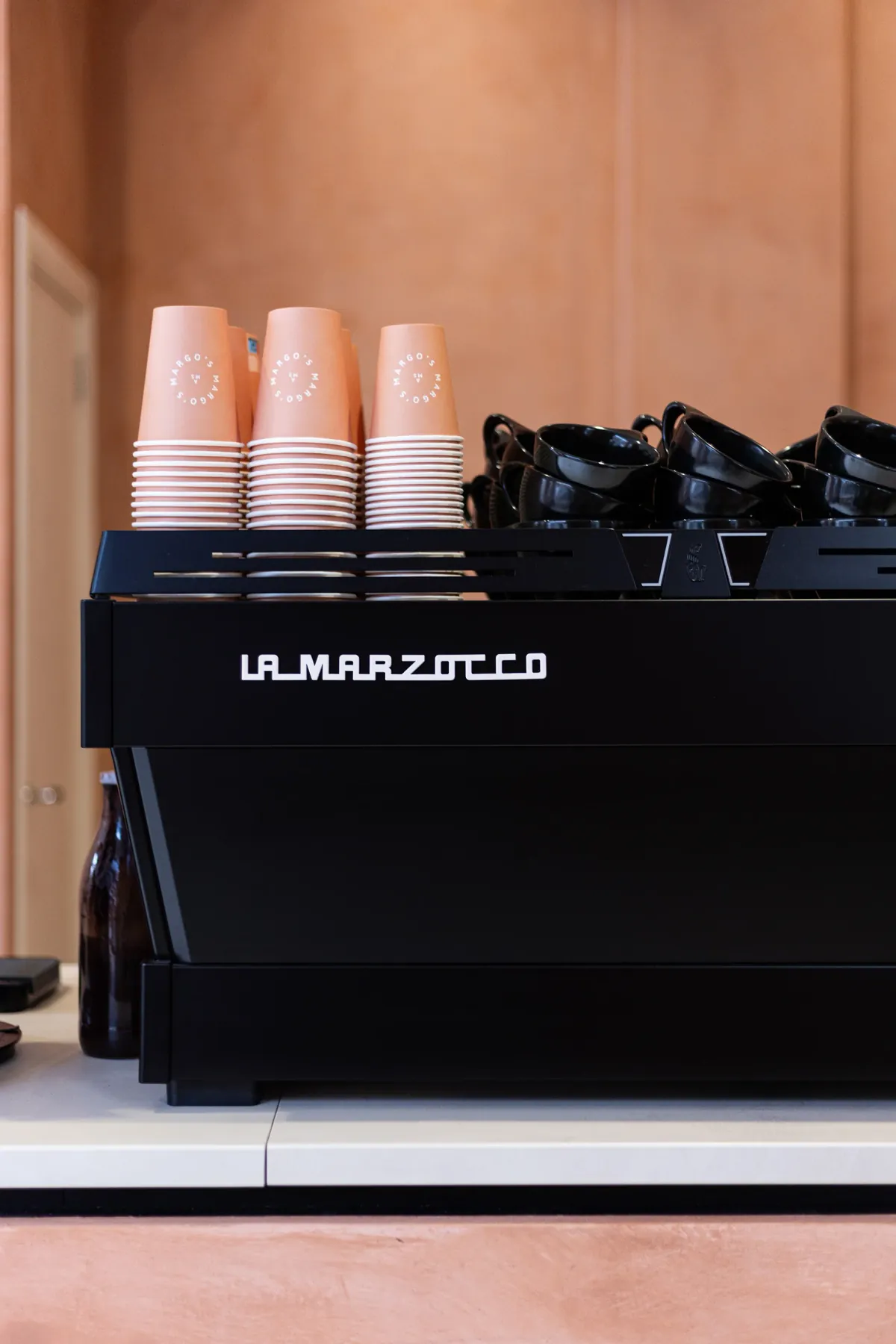 A black La Marzocco espresso machine on the counter at Margo's, with stacks of peach Margo's paper cups and black cups on top against a terracotta wall