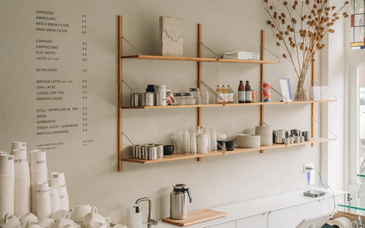 The coffee wall at SAINT-JEAN Bakery, with the drinks list lettered on a pale wall beside oak shelves of glassware, bottles and milk jugs, and paper cups stacked on the counter