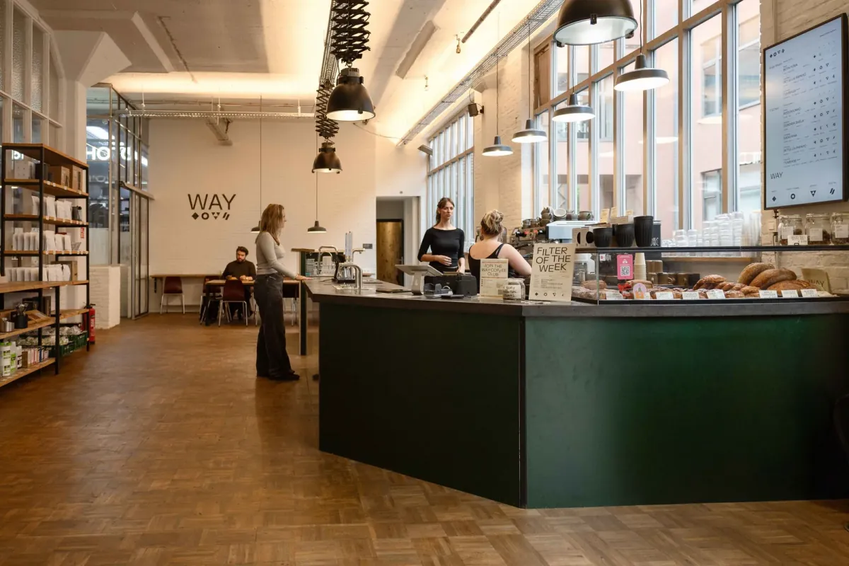The bar at WAY Dok Noord, a dark green counter with a pastry case of sourdough loaves and buns under industrial pendant lamps, tall factory windows on one side and shelves of coffee bags on the other