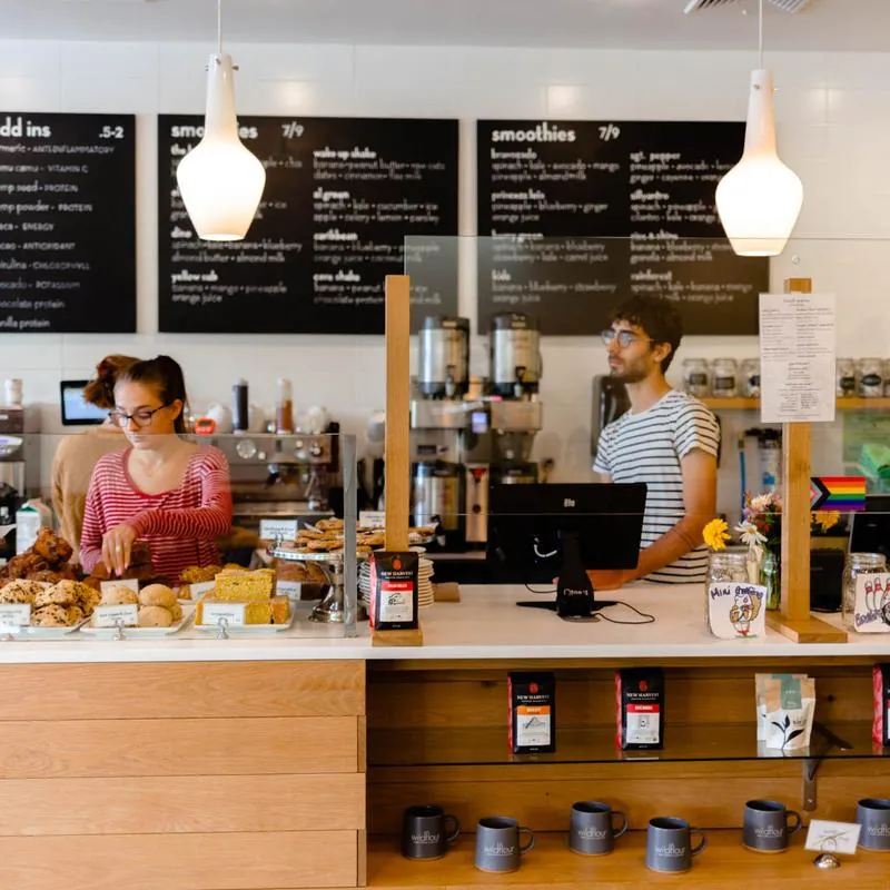 The counter at Wildflour, with pastries under glass, bags of New Harvest coffee on the shelf, branded mugs below and the smoothie boards above