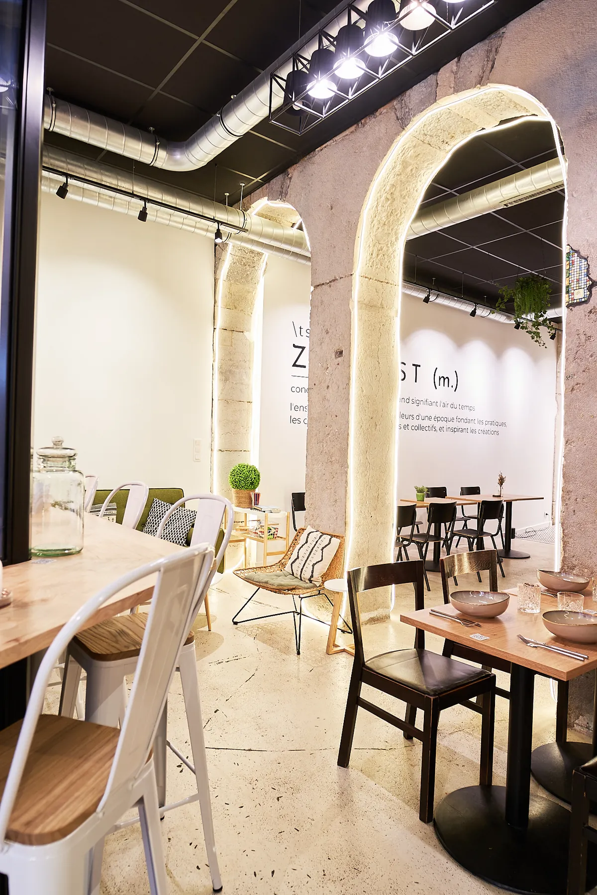 The dining room at ZEITGEIST Café in Lyon, with backlit stone arches, wooden tables, black chairs and the word Zeitgeist defined on the far wall